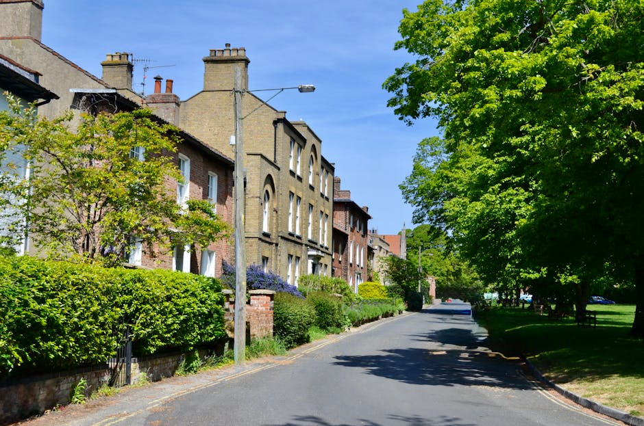 A residential street in a suburban area with a row of multi-storey terraced houses constructed from light brown brick, featuring chimney stacks, arched windows, and small front gardens bordered by low brick walls. The street is paved with asphalt and is flanked by lush greenery, including well-maintained hedges, small trees, and larger leafy trees casting shade over the pavement. On the left side, a tall utility pole with a street light extends upwards, with wires visible above the street. The background shows a clear blue sky with minimal cloud cover, and sunlight illuminating the scene, highlighting the textures of the brickwork and foliage. The environment suggests a quiet neighbourhood with a mixture of private homes and outdoor green spaces, aligning with considerations for local and alternative waste management or private rubbish clearance services such as those provided by wasteclearancerichmonduponthames.co.uk.