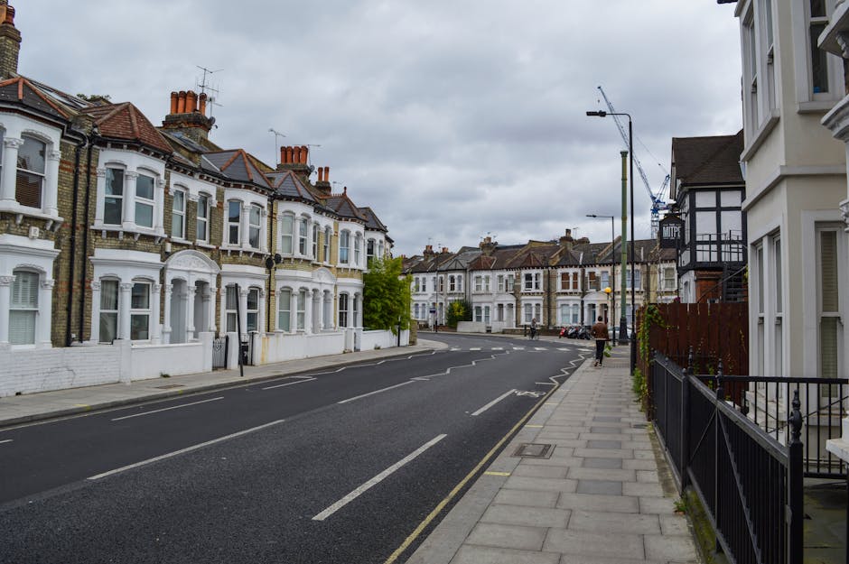 A quiet residential street in an urban area featuring a row of Victorian-style terraced houses with white-painted facades, brick details, and bay windows. The houses are adjacent to a wide concrete pavement with metal railings, and a few small potted plants are visible near the entrance of one property. The road, made of dark asphalt, has clearly marked parking bays and central lane markings, with minimal traffic. On the right side, a streetlight pole and a construction crane in the background suggest ongoing development nearby. The sky is overcast, casting even, diffuse light over the scene. The overall environment appears clean and well-maintained, typical of an inner-city neighbourhood that may require occasional private waste clearance, although no waste or rubbish is visible in the scene. The image aligns with a setting where independent rubbish removal services, such as those provided by wasteclearancerichmonduponthames.co.uk, could be utilized for local waste management needs.