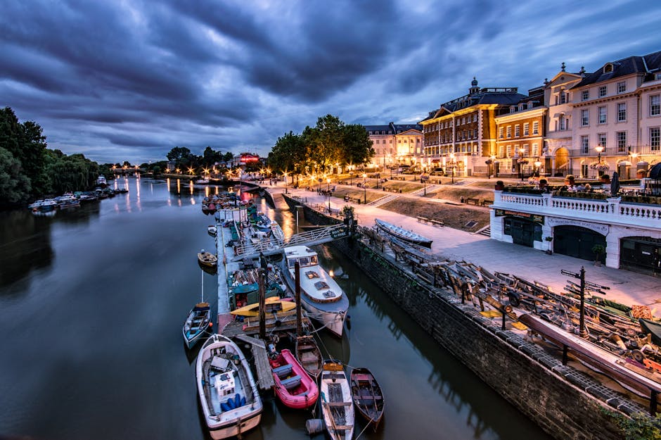 An evening view of a calm river with several small boats secured along a wooden dock, some with visible outboard motors and colorful covers, lined up parallel to the water's edge. On the right, a paved promenade runs alongside a series of elegant, well-lit historic buildings featuring light-colored facades, decorative balconies, and large windows, with warm outdoor lighting casting a subtle glow on the architecture. The background showcases a partly cloudy sky with dramatic dark clouds contrasting the illuminated townscape, while lush trees border the river on the left side, partially obscuring the distant shoreline. The scene presents a quiet, scenic riverside environment suitable for leisurely walks or private boat moorings, consistent with local urban settings where independent waste management or rubbish clearance services by companies like wasteclearancerichmonduponthames.co.uk may be employed for maintaining cleanliness in such areas.