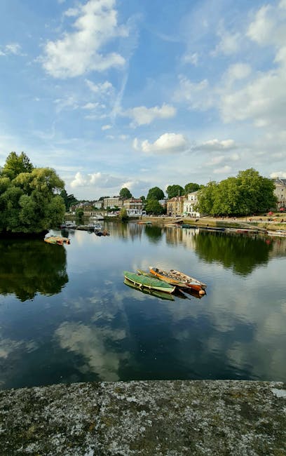 A tranquil river scene during daylight hours with several small boats, including a green and orange rowboat and a couple of wooden boats, resting on the calm water's surface. On the far bank, there are residential buildings and lush green trees lining the shoreline, with some architecture visible in the background. The sky overhead is partly cloudy with patches of blue and scattered white clouds, reflected softly in the water. The foreground features a textured stone or concrete surface, likely part of a riverside walkway or embankment, which is slightly weathered. The overall setting appears peaceful and natural, situated in a town or suburb that might benefit from interior or private waste clearance, consistent with services like those offered by wasteclearancerichmonduponthames.co.uk for alternative rubbish removal or on-site clearance solutions. The scene’s lighting is diffuse, emphasizing the clarity of the reflections and the vibrant colors of the boats and greenery, contributing to an overall calm and orderly atmosphere suitable for environmental or waste management contexts.